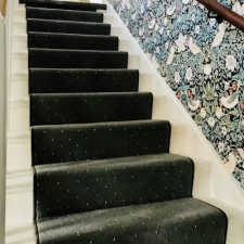 View up a staircase fitted with Adams Carpets carpet with a black whipped edge runner and a patterned wallpaper.