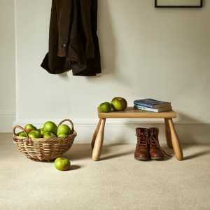 Hallway with basket of apples, footstool and coat hanging up against a neutral cream painted wall and light beige carpet in Cornish Cream from the Oaklands range