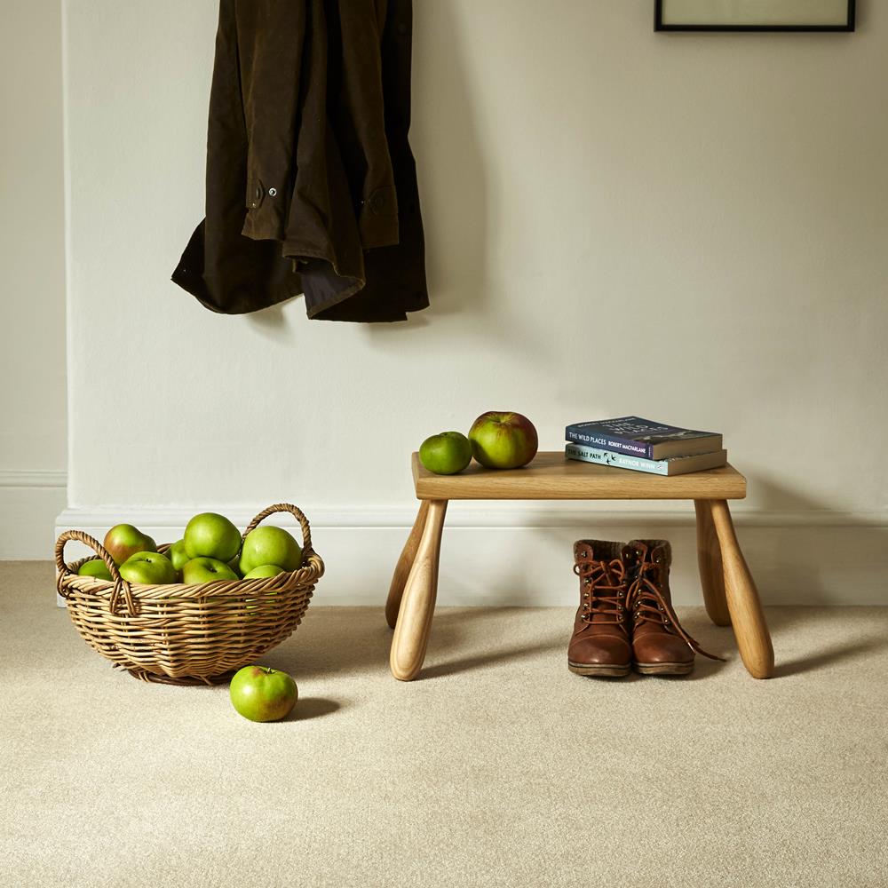 A hallway with coat hook, basket of apples and a footstool placed on an Oaklands wool twist carpet in a pale colour called Cornish Cream.