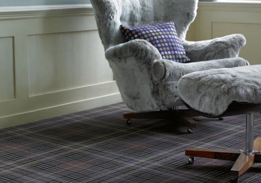 A grey armchair and footstool in a living room with a purple, black and gold plaid carpet from the Brintons Abbeyglen range.