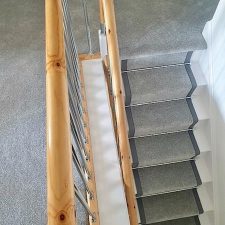View of landing and down a flight of stairs fitted with grey polypropylene carpet. The stairs have a 50mm darker grey linen binding to the stair runner and chrome effect solid stair rods.
