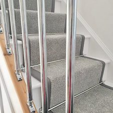 View through balustrade of stairs fitted with grey polypropylene carpet. The stairs have a 50mm darker grey linen binding to the stair runner and chrome effect solid stair rods.