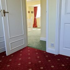 View of a room fitted with a deep red patterned carpet into a landing fitted with a green patterned woven Axminster carpet.