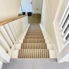View down a flight of stairs fitted with a brown and beige striped 100% wool loop carpet with brass rods to keep it in place.