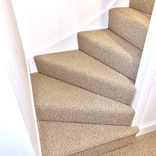 View of the bottom of a flight of stairs fitted with a beige wool loop 2 ply yarn, moth resistant carpet