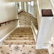View down a flight of stairs fitted with a high quality traditional woven Axminster carpet with a beige background and medallion design in red and brown.