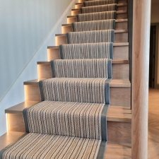 View of a flight of stairs with wooden treads and striped woven wool/nylon carpet runner with leather binding.