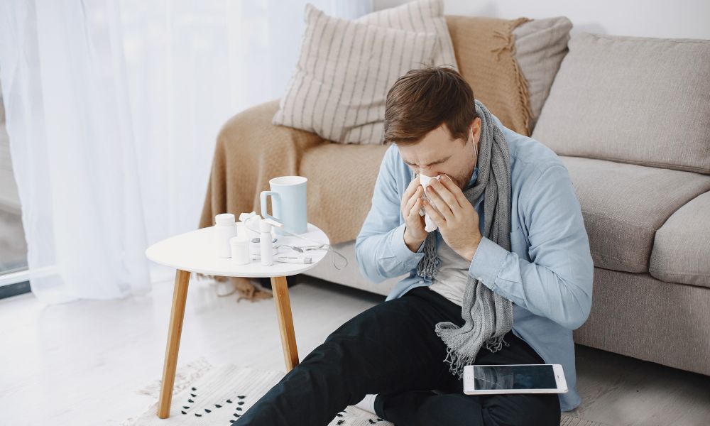 Man sitting in the floor in a living room sneezing into a tissue as he suffers from allergies and needs a hypoallergenic carpet