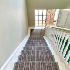 View down a flight of stairs and onto a window, the stairs are fitted with a striped runner in shades of brown, with a 40mm herringbone binding fitted to each edge.