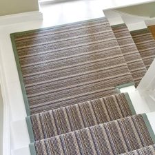 View down a flight of stairs and onto a landing half way down fitted with a striped runner in shades of brown, with a 40mm herringbone binding fitted to each edge.