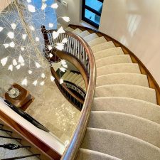 View down a curved staircase with wooden balustrade and grey wool carpet which has been fully stuck and stapled.