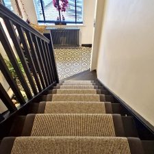View down a flight of stairs fitted with Ceneva Actionbac carpet runner with mid grey herringbone binding and solid brass stair rods coated in matt black finish.