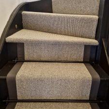 View of a flight of stairs fitted with Ceneva Actionbac carpet runner with mid grey herringbone binding and solid brass stair rods coated in matt black finish.