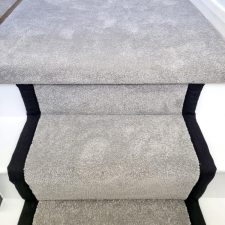 View of the top of a flight of stairs and onto an upstairs landing, covered with a grey twist pile, bleach cleanable carpet and herringbone binding to the sides.