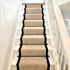 View up a flight of stairs fitted with a light beige polypropylene carpet stair runner with black herringbone binding and black stair rods.