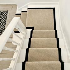 View down a flight of stairs fitted with a light beige polypropylene carpet stair runner with black herringbone binding and black stair rods.