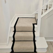 The bottom of a flight of stairs fitted with a polypropylene carpet stair runner with black herringbone binding and black stair rods.