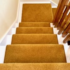 View down a flight of stairs fitted with a wool twist carpet runner in a mustard colour with white painted stairs and skirting board.