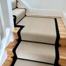 View of a landing with flight of stairs fitted with a herringbone-patterned cream carpet with black binding and black rods.